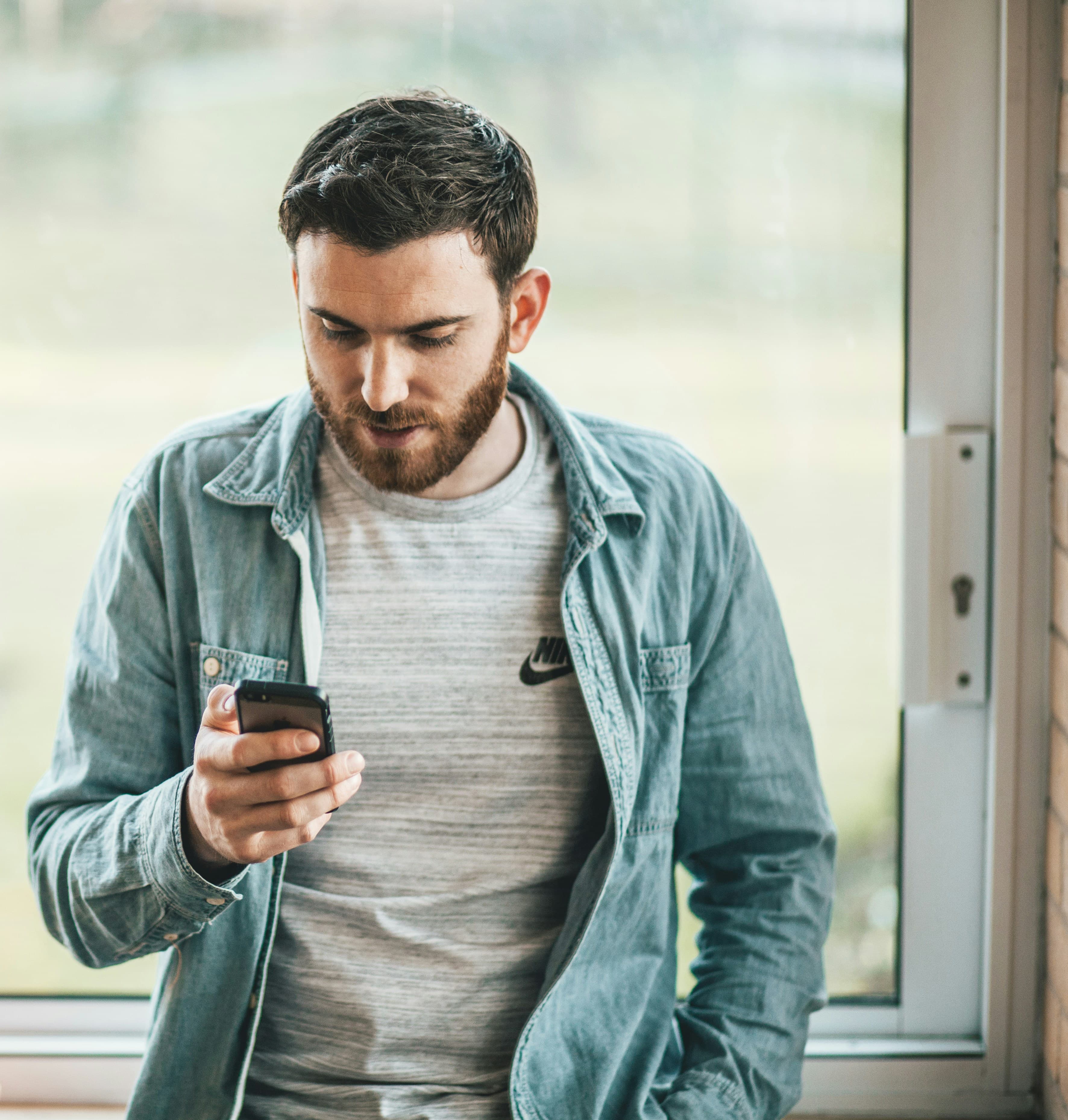 A man looking at his phone beside a window.
